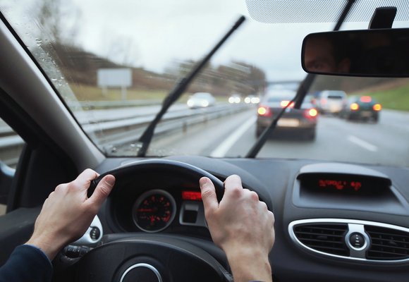 Dopuszczalne pęknięcie szyby przedniej w samochodzie Dopuszczalne pęknięcie szyby przedniej w samochodzie, widok z wnętrza pojazdu z rękami na kierownicy, deszczowa pogoda, w tle ruch uliczny.