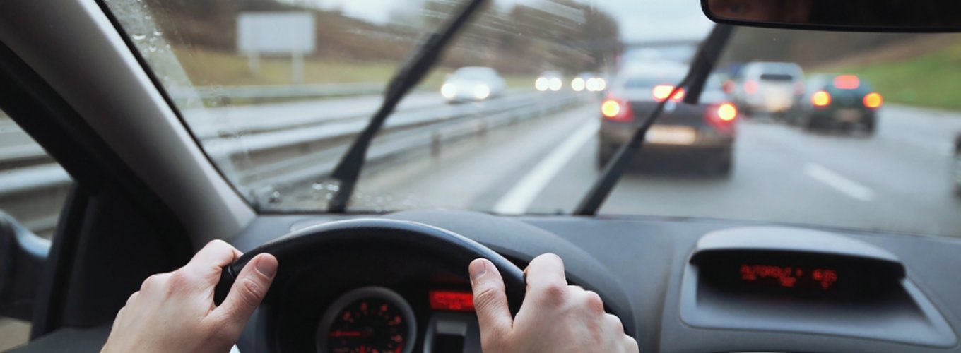 Dopuszczalne pęknięcie szyby przedniej w samochodzie, widok z wnętrza pojazdu z rękami na kierownicy, deszczowa pogoda, w tle ruch uliczny.