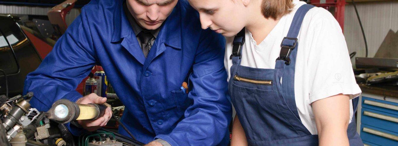 Dwóch mechaników pracuje nad silnikiem samochodu w warsztacie. Mężczyzna w niebieskim kombinezonie używa latarki, wskazując na detal silnika, podczas gdy kobieta w białej koszuli i niebieskim fartuchu uważnie obserwuje.
