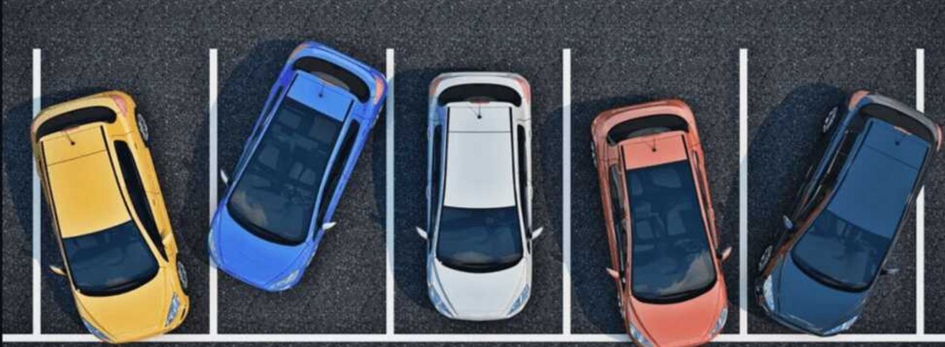 Cztery samochody zaparkowane na parkingu, jeden z nich - niebieski pojazd - zajmuje dwa miejsca parkingowe, co może skutkować mandatem za nieprawidłowe parkowanie.