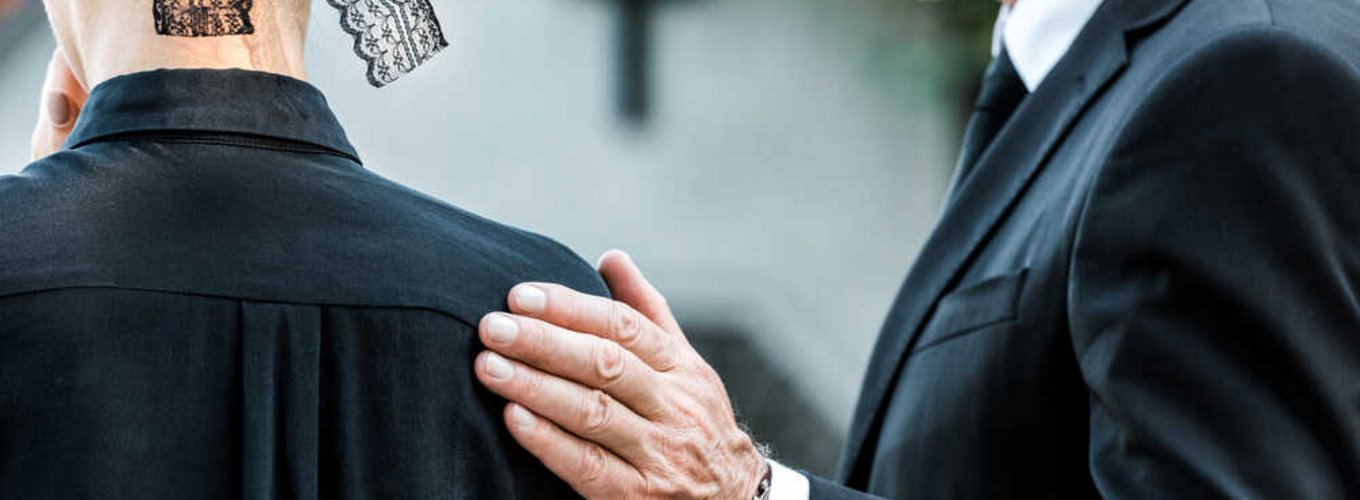 Dwie osoby ubrane w żałobne stroje stoją odwrócone plecami. Mężczyzna trzyma dłoń na ramieniu kobiety i pocieszą ją.