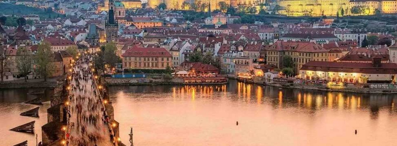 Widok na zachodzące słońce nad Pragą z widocznym Zamkiem Praskim i katedrą św. Wita w oddali, a na pierwszym planie tłum ludzi przechodzących przez kamienny Most Karola nad rzeką Wełtawą.