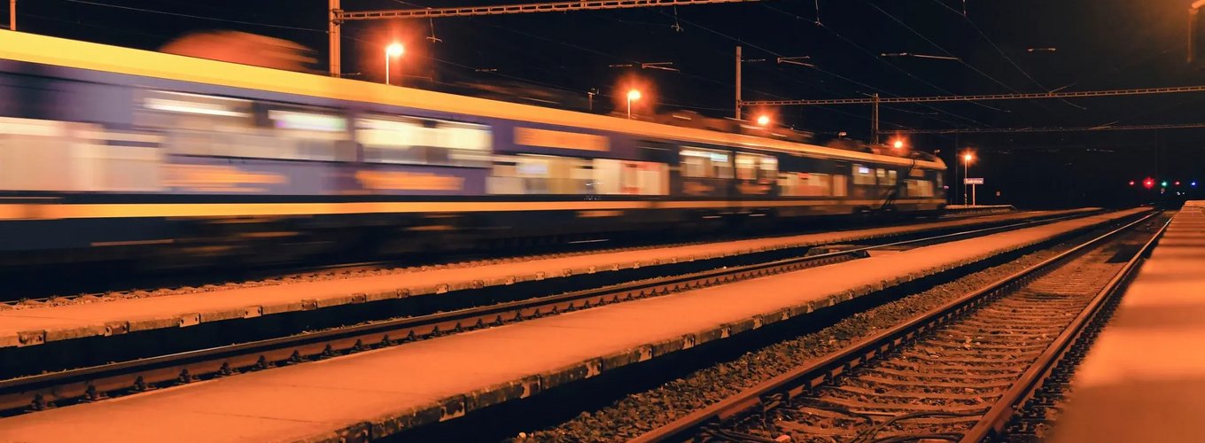 Pędzący pociąg na torach kolejowych nocą z rozmazanym efektem ruchu, oświetlony ulicznymi latarniami, z widoczną siecią trakcyjną i sygnalizacją świetlną w tle.