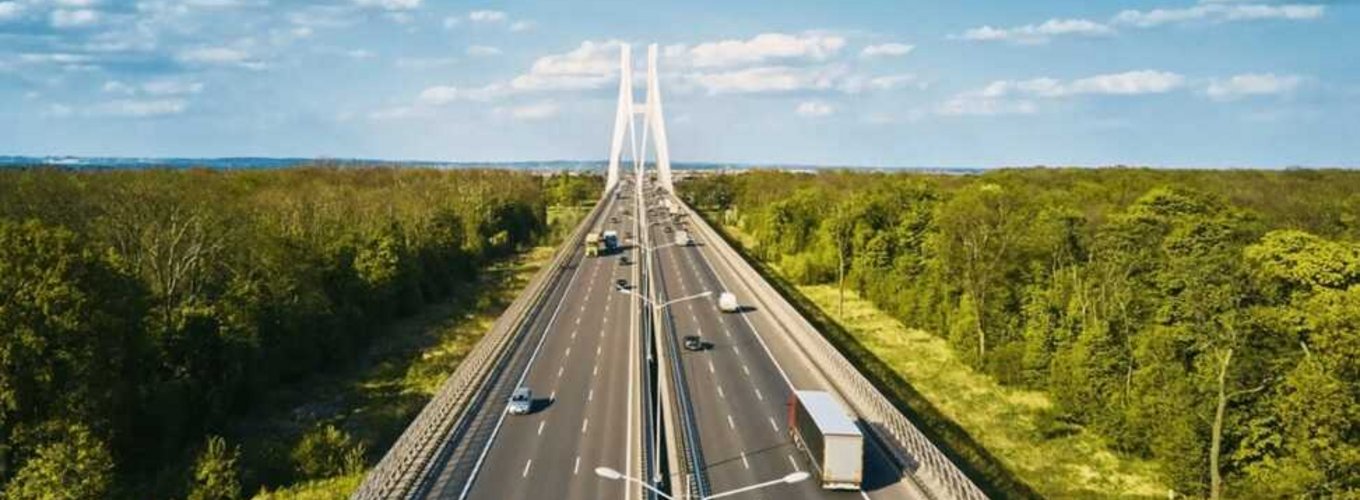 Widok na nowoczesny wielopasmowy autostradowy most z białymi kablami, po którym poruszają się różne pojazdy w obu kierunkach, z ciężarówką i samochodami osobowymi – potencjalny przykład drogi objętej systemem opłat drogowych eurowinieta