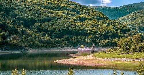 Spokojny krajobraz z zielonymi wzgórzami otaczającymi spokojny zbiornik wodny, w którym znajduje się częściowo zanurzony stary budynek, przypominający kościół.