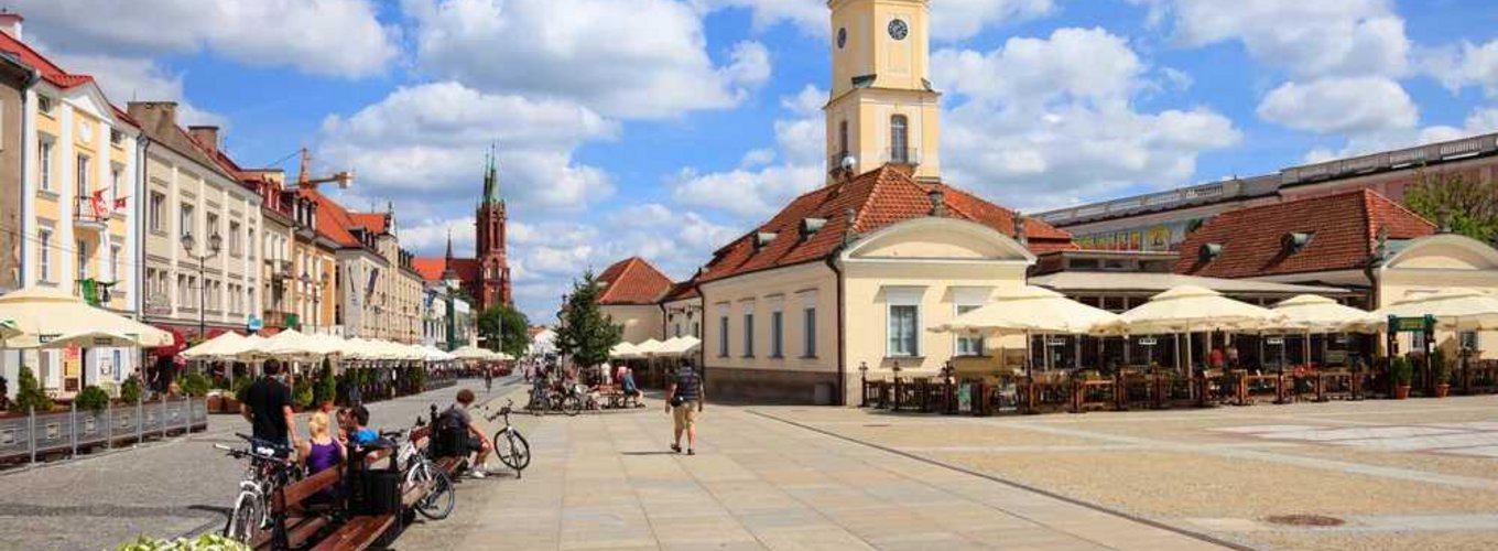 Widok na malowniczy, słoneczny rynek w Białymstoku z widocznym zegarem na wieży, idealne miejsce na spacer z kartą EKUZ w kieszeni. Na pierwszym planie ławki z siedzącymi ludźmi, rowery oraz kwiaty, a w tle restauracje z parasolami.