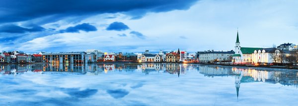 Panoramiczny widok na miasteczko z kolorowymi budynkami i kościołem z zieloną wieżą, odbijający się w spokojnej wodzie pod dramatycznym, pochmurnym niebem