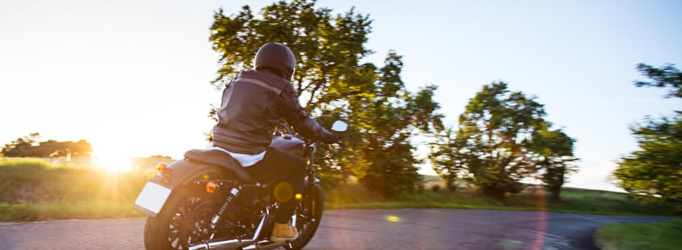 Osoba ubrana w kask i kurtkę jedzie motocyklem po wiejskiej drodze o zachodzie słońca.