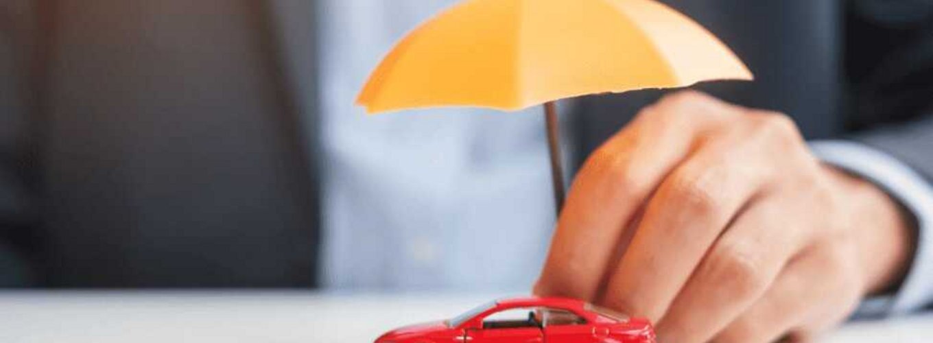 Miniaturka czerwonego samochodu chroniona przez pomarańczową miniaturową parasolkę trzymaną przez dłoń osoby w eleganckim garniturze, symbolizująca ubezpieczenie OC krótkoterminowe dla pojazdów.