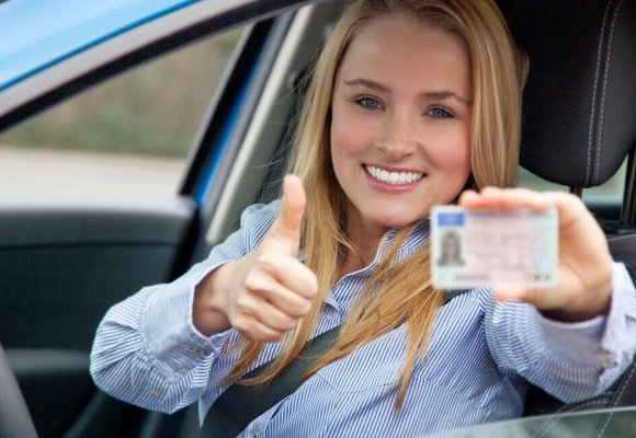 Młoda kobieta siedzi w aucie i przez otwarte okno z uśmiechem prezentuje prawo jazdy kategorii B oraz uniesiony w górę kciuk