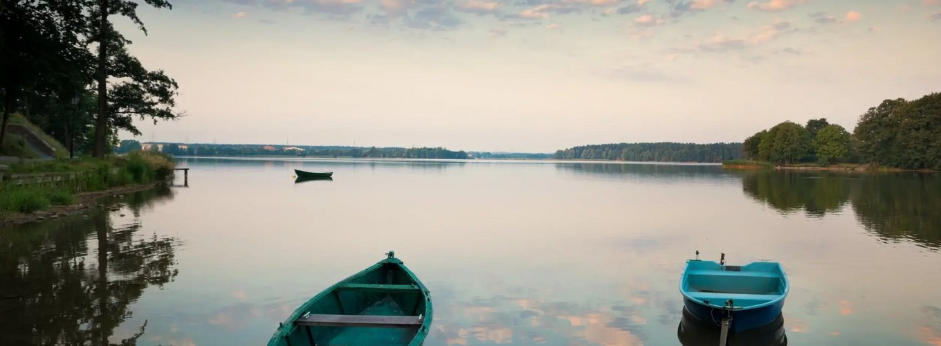 Spokojne jezioro o zachodzie słońca z dwiema łodziami na pierwszym planie, niebo z delikatnymi chmurami odbijającymi się w wodzie, linia brzegowa z drzewami.