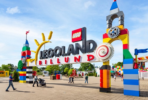 Wejście do LEGOLAND Billund z kolorową bramą z klocków LEGO, ozdobioną różnymi dekoracjami LEGO, takimi jak słońce, koła zębate i wieże, z napisem "LEGOLAND BILLUND" na górze.