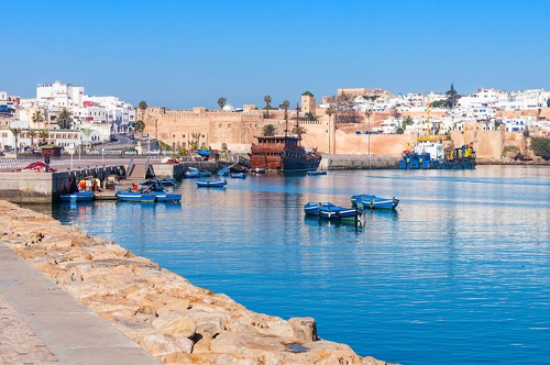 Medina w Rabacie Na zdjęciu widać malowniczy widok na port z kilkoma niebieskimi łodziami unoszącymi się na spokojnej wodzie, kamiennym chodnikiem na pierwszym planie oraz historycznym miastem z białymi budynkami i palmami w tle.