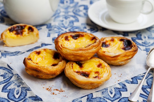 Cztery portugalskie tarty z kremem (pastéis de nata) leżą na pergaminie, w tle biały imbryk i filiżanka, wszystko na niebiesko-białym wzorzystym obrusie.