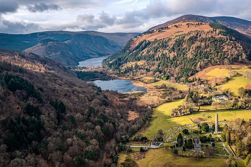 Wicklow National Park, Ireland Widok z lotu ptaka na malowniczą dolinę z jeziorem, otoczoną zalesionymi wzgórzami i górami, z rozproszonymi budynkami i cmentarzem na pierwszym planie.