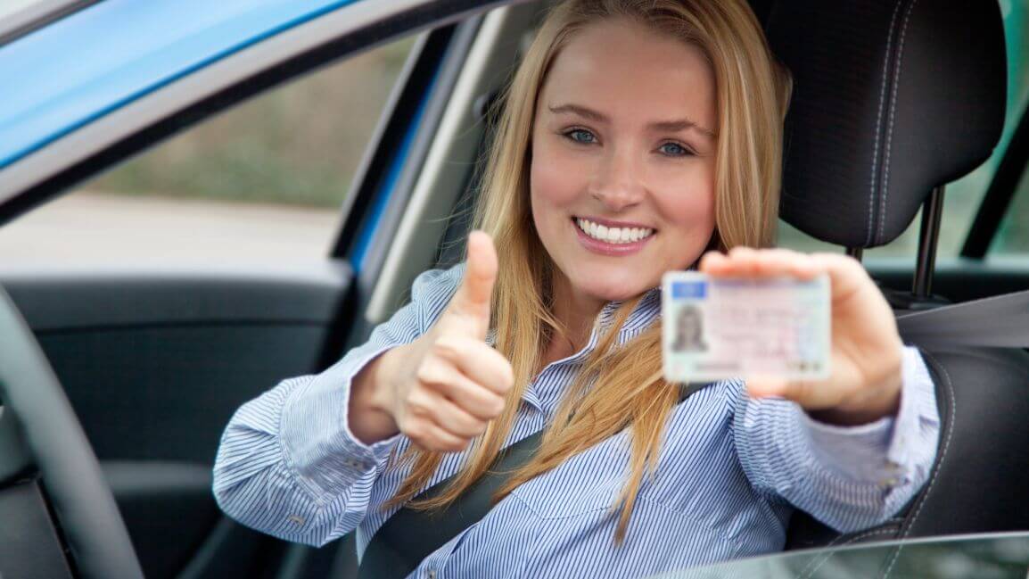 Młoda kobieta siedzi w aucie i przez otwarte okno z uśmiechem prezentuje prawo jazdy kategorii B oraz uniesiony w górę kciuk