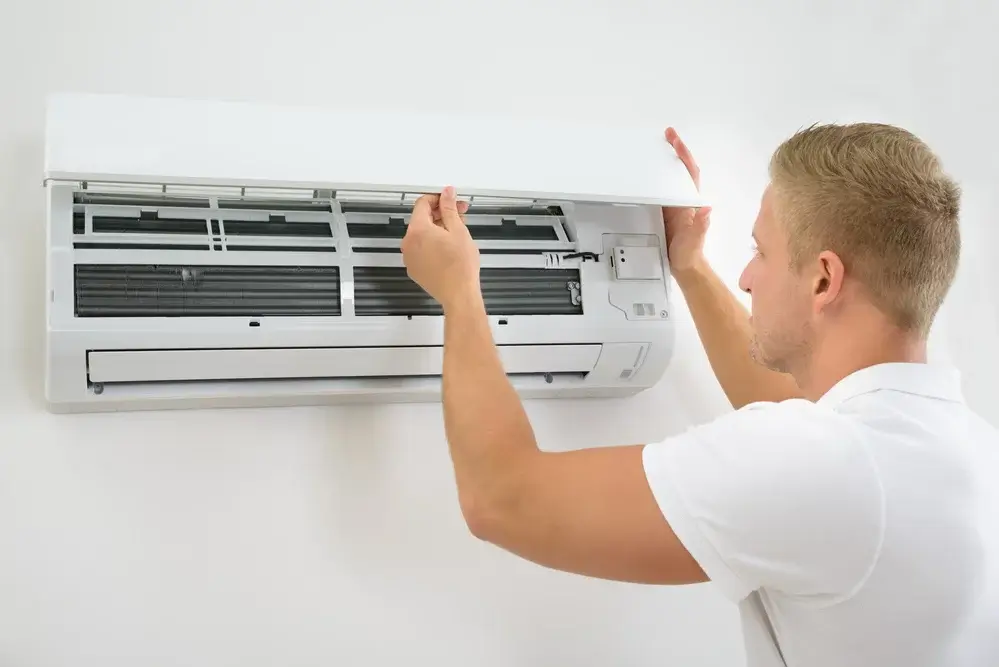 Mężczyzna w białej koszulce serwisuje klimatyzację w mieszkaniu, sprawdzając filtr i wewnętrzne komponenty, zapewniając utrzymanie optymalnej temperatury i jakości powietrza w pomieszczeniu.